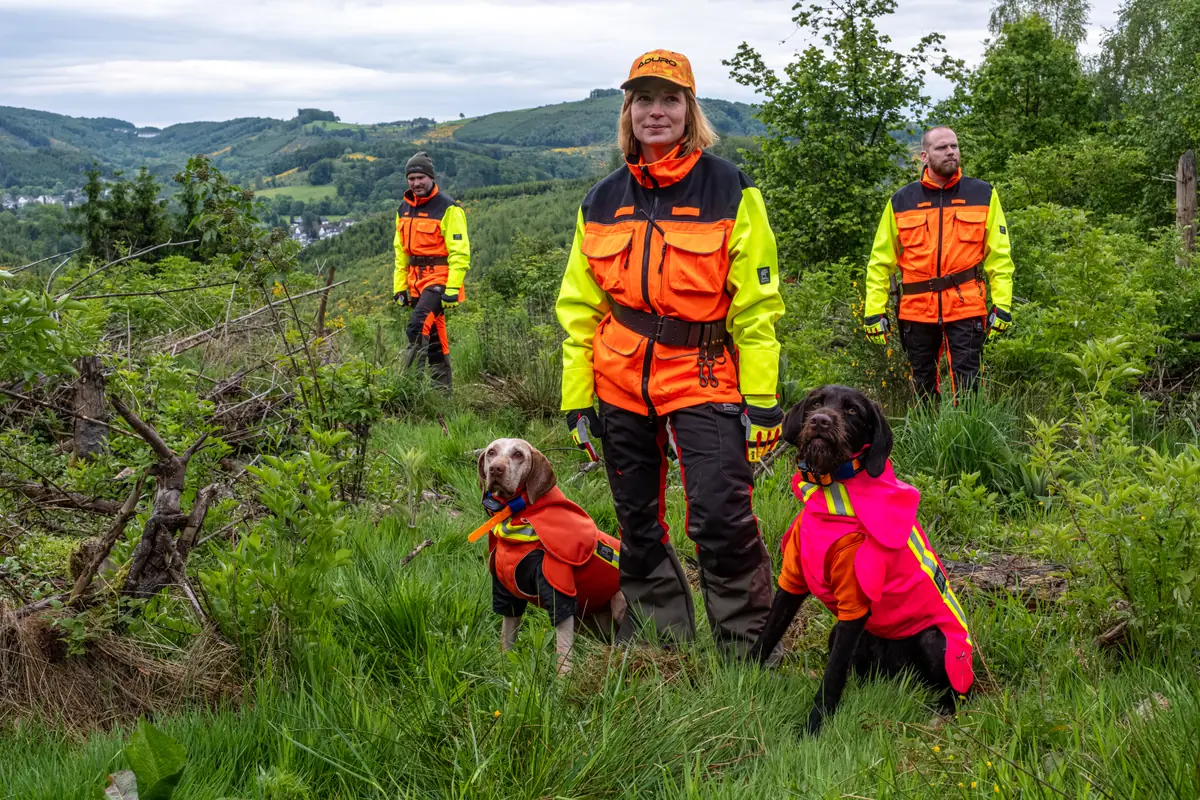 Drei Hundeführer mit zwei Jagdhunden in ADURO Schutzwesten (Orange und Pink) im Revier